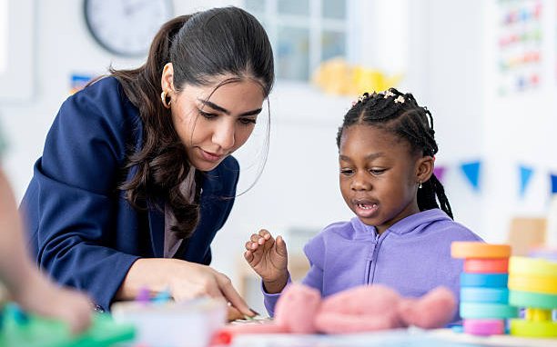 A teacher kneeling beside a young girl to help her with an activity at a classroom table