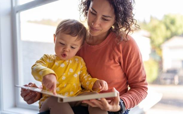 A caregiver holding a baby on her lap while the baby points at a page in a picture book
