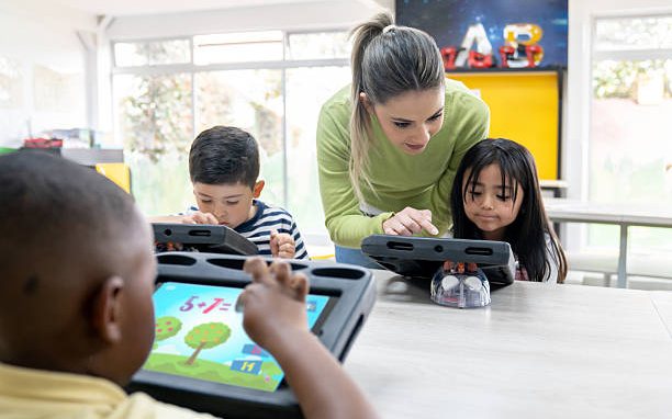A teacher leaning over a classroom table to help three preschoolers work on learning tablets